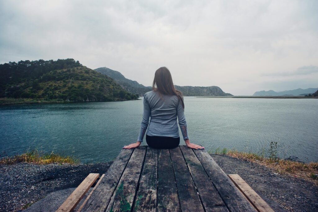 Woman Wearing Gray Long-sleeved Shirt and Black Black Bottoms Outfit Sitting on Gray Wooden Picnic Table Facing Towards Calm Body of Water at Daytime