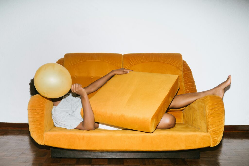 A person lies on a yellow sofa, holding a balloon with a playful, relaxed pose indoors.