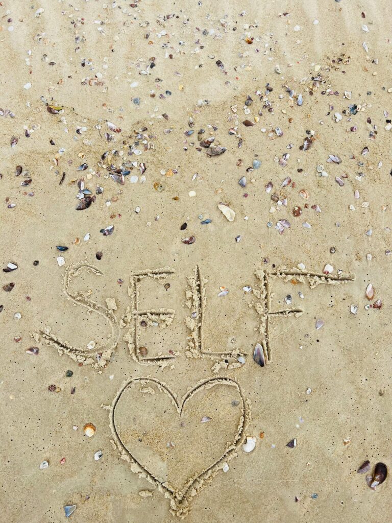 The word 'self' with a heart shape drawn in sand amidst scattered seashells on a beach.