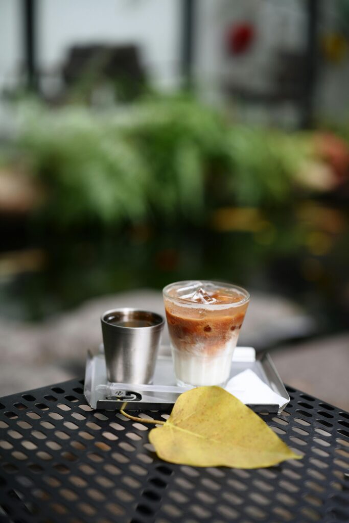 A glass of iced coffee with metal cup on a garden table with a yellow leaf, set outdoors.