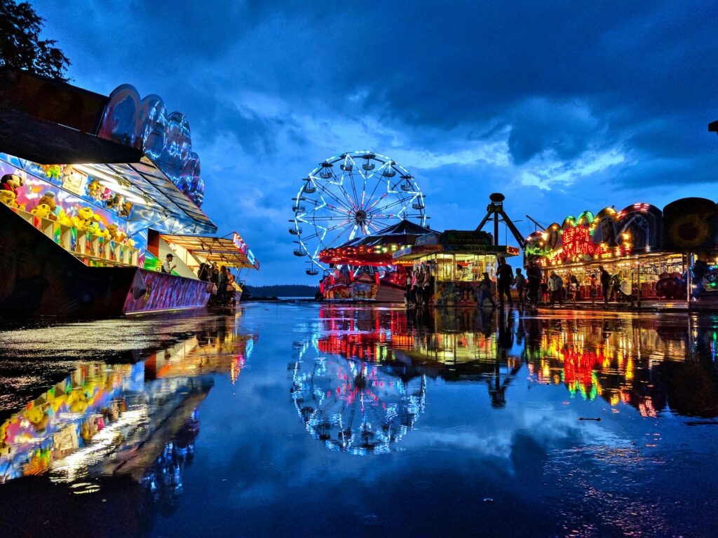 A colorful carnival at dusk reflecting on wet ground with a Ferris wheel and bright lights.