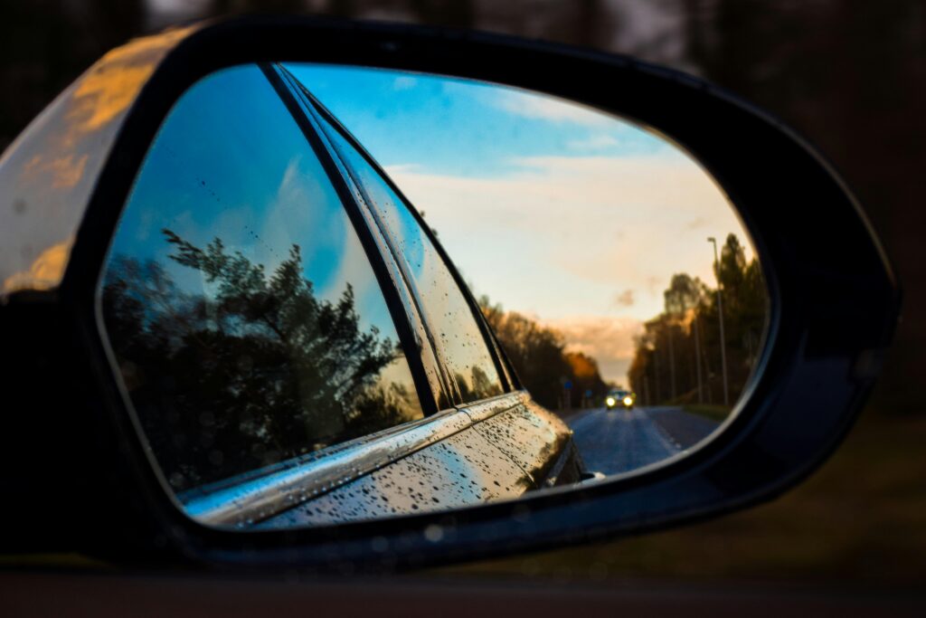 A wet side mirror capturing a dynamic reflection of a road and trees at twilight.