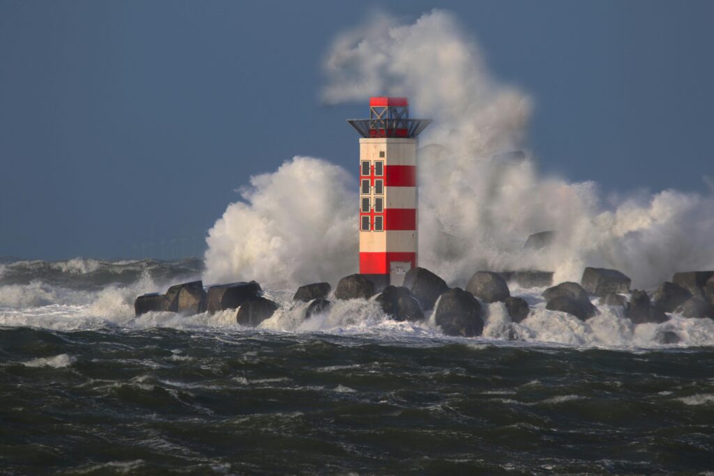 pexels photo 3722772 3722772 Red and white lighthouse with waves crashing against rocks under a stormy sky.