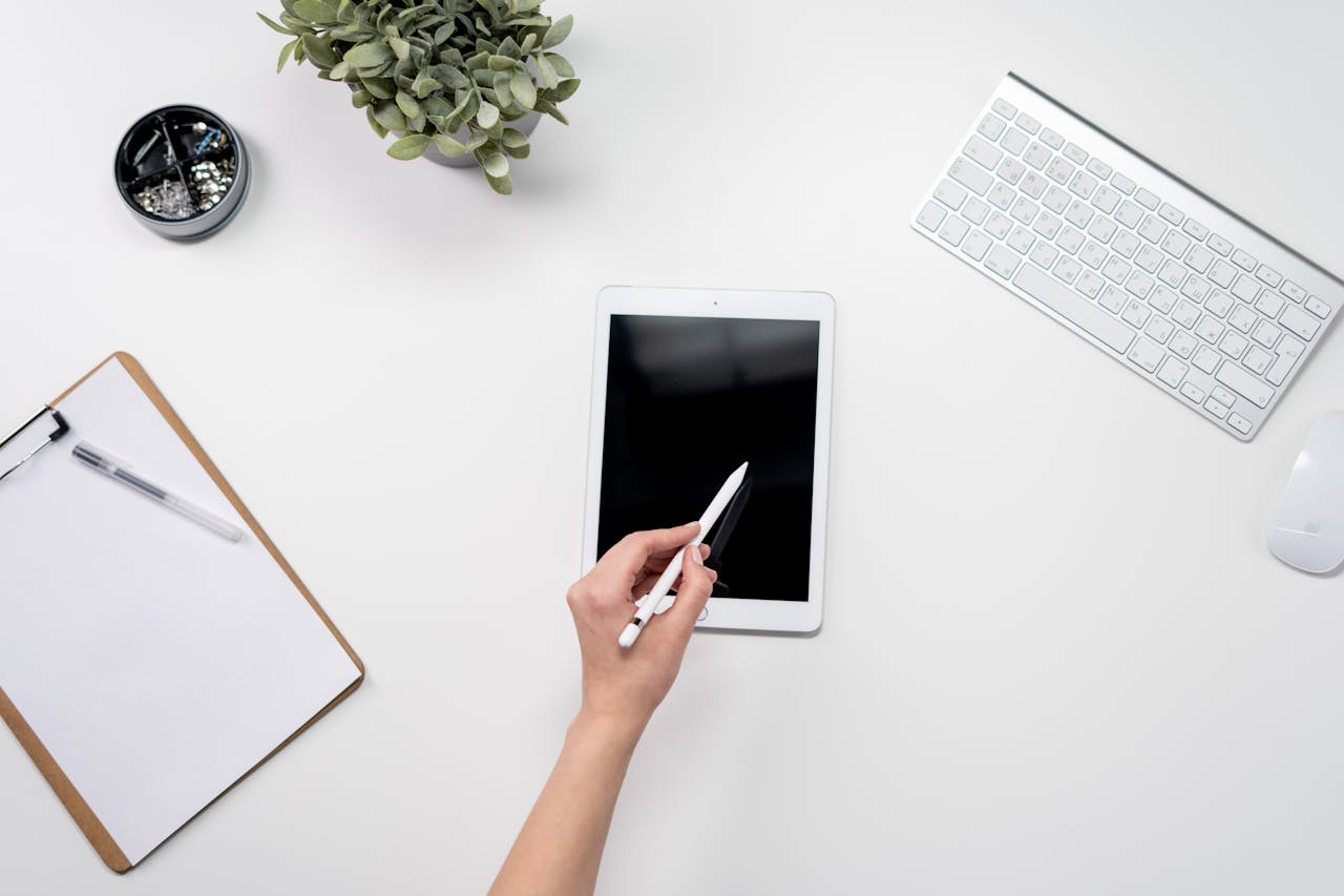 A minimalist home office setup featuring a tablet, stylus, keyboard, plant, and stationery on a white desk.