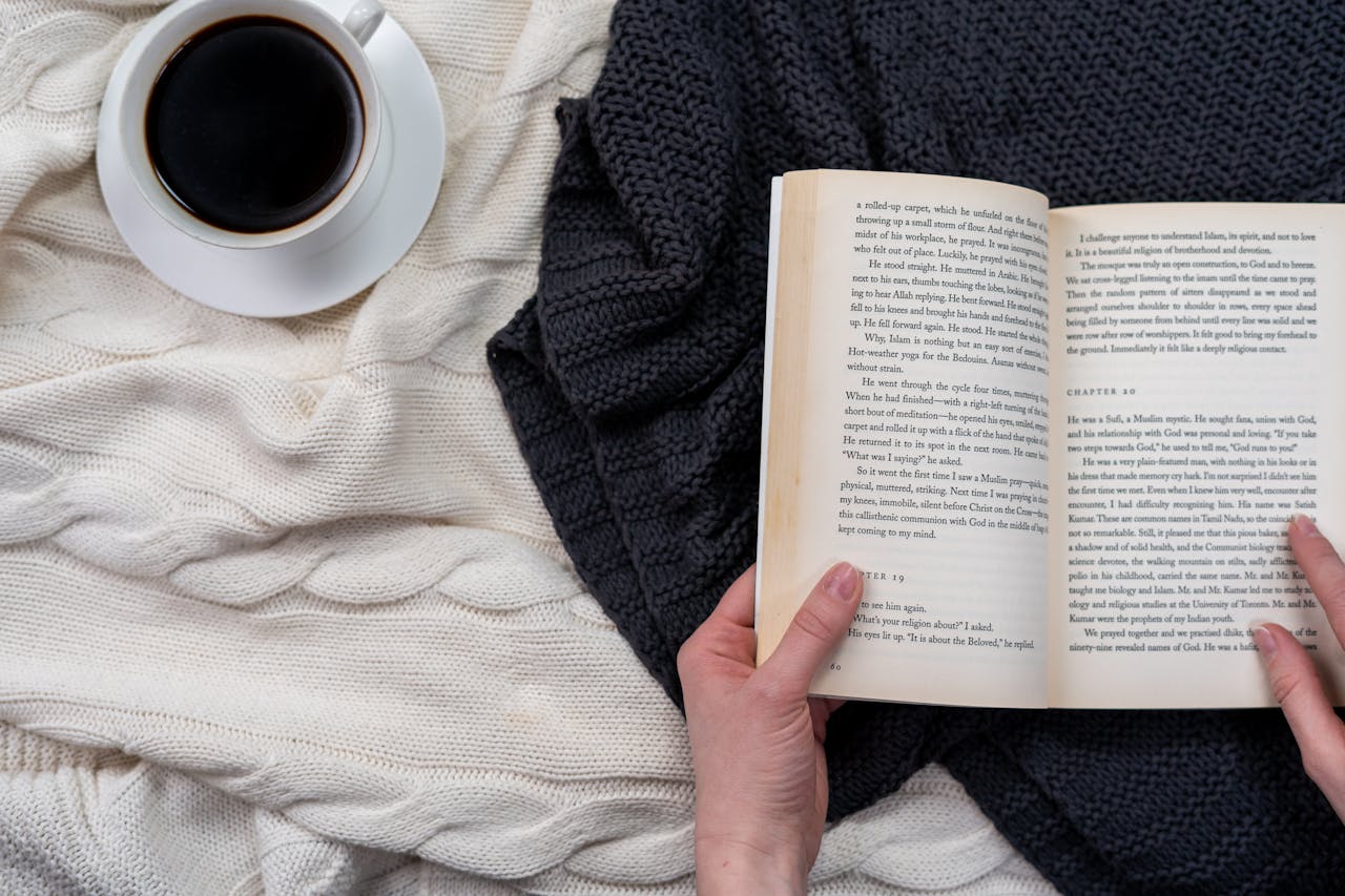 Overhead view of hands holding a book beside a coffee cup on a cozy blanket.